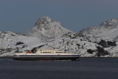 Bodo, Norveç - 4 Ocak 2023. Arka planda karla kaplı dağlar olan bir feribotun telefoto görüntüsü. Bunun gibi feribotlar Norveç toplu taşıma sisteminde önemli bir rol oynamaktadır.