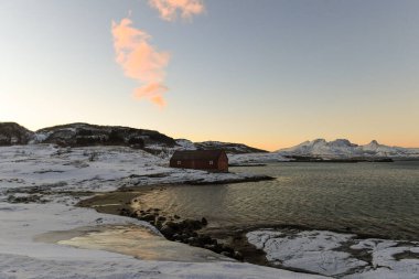 A traditional norwegian red barn enclosed by rugged mountains and snow-covered beaches in the arctic. Shown during the golden hour of a brief period of daylight near the town of Bodo. clipart