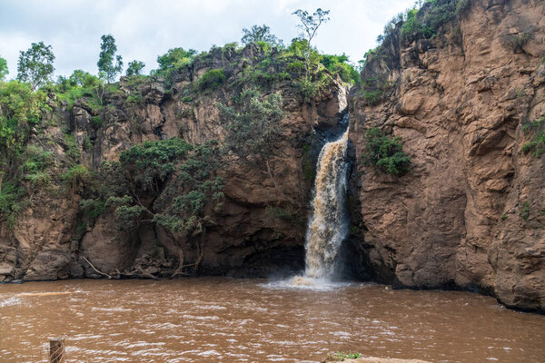 Malika Falls in Nakuru National Park, Kenya, is a stunning waterfall cascading down rocky cliffs surrounded by lush greenery. The water flows into a small pool below, creating mist and a soothing
