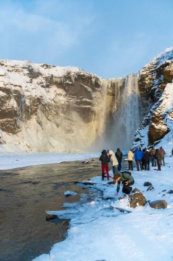 Skogafoss, İzlanda - 2 Ocak 2025. Karlı bir arazide kısmen donmuş bir şelaleyi izleyen turistler. Bu sonbahar, İzlanda 'nın güney kıyıları boyunca en popüler yerlerden biri oldu.