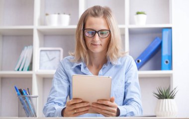 Bir sandalyede oturan ve çalıştığı yerdeki tablet üzerinde çalışan genç ve güzel bir iş kadını. Ofis işi kavramı