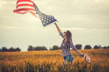 Kolza tohumu tarlasında rüzgarda Amerikan bayrağını tutan güzel bir genç kız. Mavi gökyüzüne karşı yaz manzarası. Bağımsızlık Günü 4 Temmuz