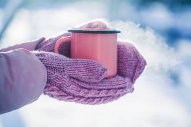 Bulanık bir kar manzarasının arka planına karşı örgü eldivenler giyenlerin elinde kırmızı bir kupa. Kış sabahı için ılık bir içecek.