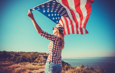 Güzel bir genç kız, çayırda rüzgarda Amerikan bayrağı taşıyor. Mavi gökyüzüne karşı yaz manzarası. Bağımsızlık Günü 4 Temmuz