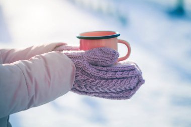 a red mug in the hands of those dressed in knitted mittens against the backdrop of a blurred snow landscape. a warming drink for a winter morning clipart