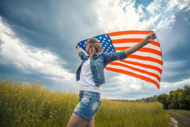 Kolza tohumu tarlasında rüzgarda Amerikan bayrağını tutan güzel bir genç kız. Mavi gökyüzüne karşı yaz manzarası. Bağımsızlık Günü 4 Temmuz