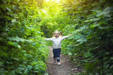 Gülümseyen kız yaprak döken ormanda yolda koşar. El sallıyor. Parlak ışık arkaplanındaki orman.