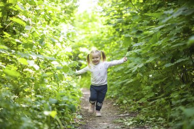 Gülümseyen kız yaprak döken ormanda yolda koşar. El sallıyor. Parlak ışık arkaplanındaki orman.