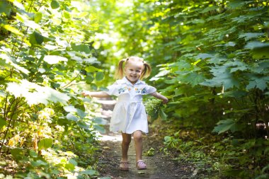 Beyaz elbiseli gülümseyen kız yaprak döken ormanda yolda koşuyor. El sallıyor. Parlak ışık arkaplanındaki orman.