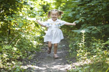 Beyaz elbiseli gülümseyen kız yaprak döken ormanda yolda koşuyor. El sallıyor. Parlak ışık arkaplanındaki orman.