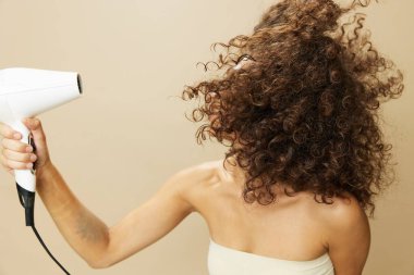 Kadın saç kurutma makinesiyle kıvırcık saç kurutuyor, ev stil ürünlerini yeniyor, bej arka planda gülümsüyor. Yüksek kalite fotoğraf