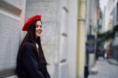 Şehirde bir duvarın yanında duran şık bir ceket ve kırmızı dudaklı kırmızı bereli bir kadın seyahat ediyor. Yüksek kalite fotoğraf