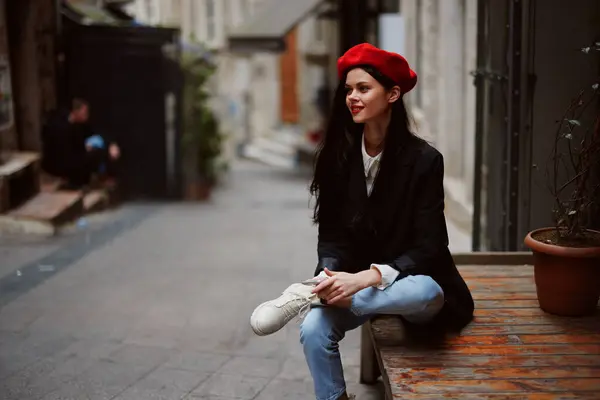 Stylish woman tourist in stylish clothes in a jacket and red beret sitting outside with phone in hand, cell phone in travel, French style, cinematic color. High quality photo
