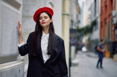 Şehirde bir duvarın yanında dikilen bir kadın şık bir ceket ve kırmızı dudaklı kırmızı bereli bir kadın seyahat ve boş zaman, Fransız tarzı elbise, bahar. Yüksek kalite fotoğraf