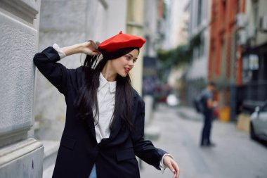 Şehirde bir duvarın yanında dikilen bir kadın şık bir ceket ve kırmızı dudaklı kırmızı bereli, seyahat ve boş zaman, Fransız tarzı bir elbise giyiyor. Yüksek kalite fotoğraf