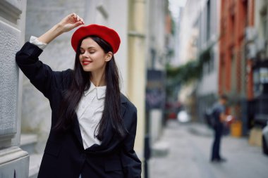 Şehirde bir duvarın yanında dikilen bir kadın şık bir ceket ve kırmızı dudaklı kırmızı bereli, seyahat ve boş zaman, Fransız tarzı bir elbise giyiyor. Yüksek kalite fotoğraf