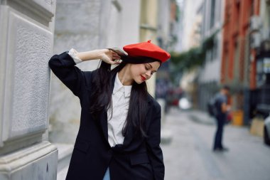 Şehirde bir duvarın yanında dikilen bir kadın şık bir ceket ve kırmızı dudaklı kırmızı bereli, seyahat ve boş zaman, Fransız tarzı bir elbise giyiyor. Yüksek kalite fotoğraf