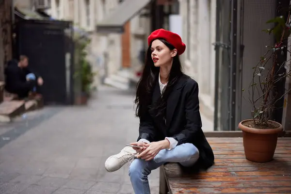 Stylish woman tourist in stylish clothes in a jacket and red beret sitting outside with phone in hand, cell phone in travel, French style, cinematic color. High quality photo
