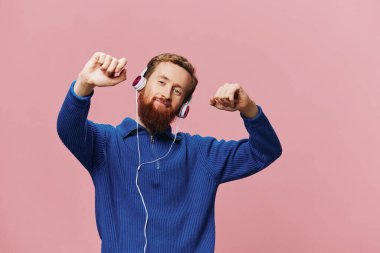 Kulaklık takan, gülümseyen ve dans eden, pembe arka planda müzik dinleyen kızıl saçlı bir adamın portresi. Sakallı bir hippi. Yüksek kalite fotoğraf