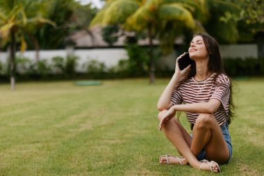 Avuç içi kadını mobil çim çizgili beyaz tenli, hayat tarzını kullanan mutlu kot pantolon çantası gülümseyen ağaç blogcusu telefon parkında gülümseyen zeki.
