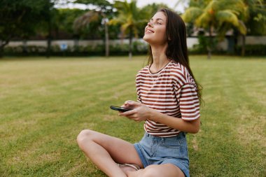 Uygulamalı kadın dışarıda gülümseyen parkta konuşuyor zeki yaz mutlu yaşam tarzı tişörtlü kız Palm Doğa Telefon Çim Ağacı blogcusunu arıyor