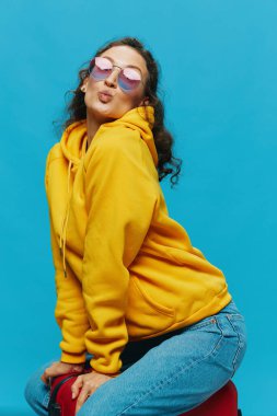 Sarı kapüşonlu bir çantada oturan gülümseyen kadın mavi arka planda mavi kot pantolon ve gözlüklü, yolculuk için hazırlanıyor. Yüksek kalite fotoğraf