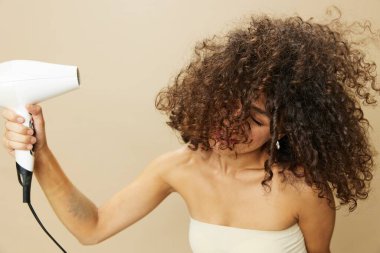 Kadın saç kurutma makinesiyle kıvırcık saç kurutuyor, ev stil ürünlerini yeniyor, bej arka planda gülümsüyor. Yüksek kalite fotoğraf