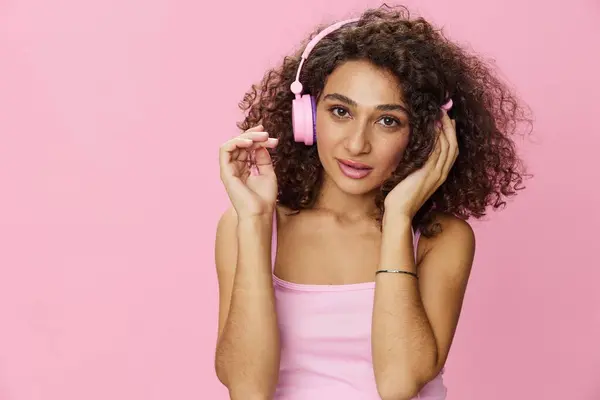 Happy woman wearing headphones with curly hair listening to music and singing along with her eyes closed in a pink T-shirt and jeans on a pink background DJ party, copy space. High quality photo