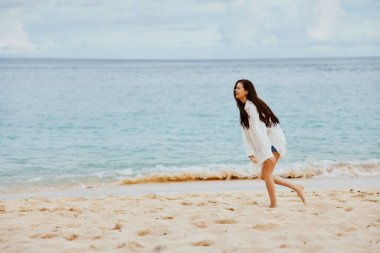 Spor kadın kumsalda koşuyor. Sarı tişört ve kot şortla kumsalda koşuyor. Uçan beyaz gömlek, okyanus manzarası, plaj tatili. Yüksek kalite fotoğraf
