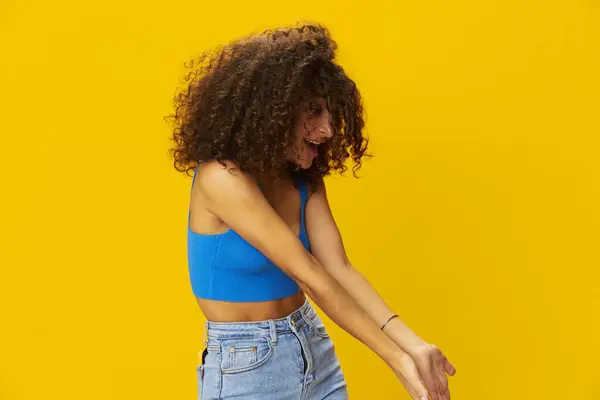 Woman with curly afro hair in a blue t-shirt on. yellow background signs with her hands, look into the camera, smile with teeth and happiness, copy space. High quality photo