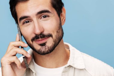 Telefonda konuşan esmer bir adamın yakın plan portresi, dişleri açık bir gülümseme ağız mutluluğu, beyaz bir tişört ve kot pantolon içinde mavi bir arka plan, kopyalama alanı. Yüksek kalite fotoğraf