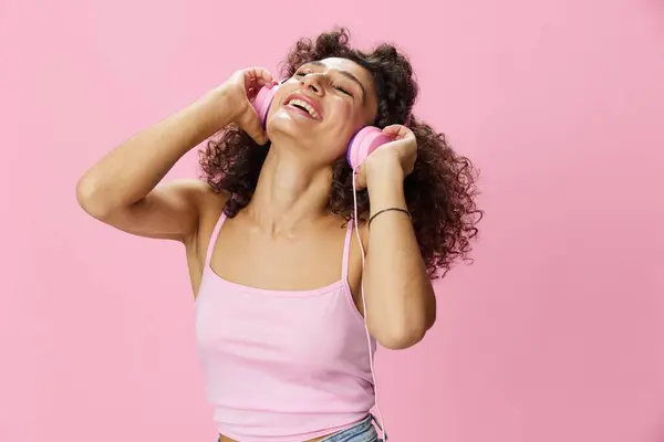 Happy woman wearing headphones with curly hair listening to music and dancing in a pink T-shirt and jeans on a pink background, copy space. High quality photo