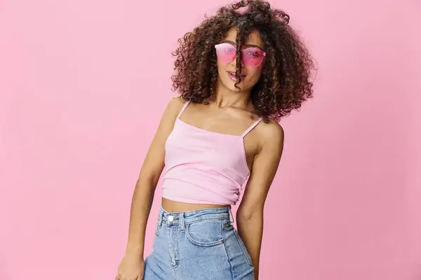 Happy woman afro curls hair dancing on a pink background in summer pink t-shirt jeans and glasses, summer vibe, copy space. High quality photo