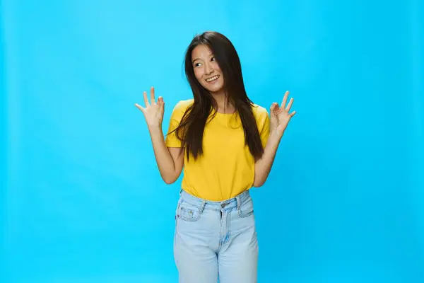 青い背景に黄色いTシャツと青いジーンズの緩い背景で指を指して歯で微笑むアジアの女性 コピースペース 高品質の写真 — ストック写真