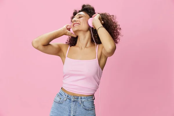 Happy woman wearing headphones with curly hair listening to music and dancing in a pink T-shirt and jeans on a pink background, copy space. High quality photo