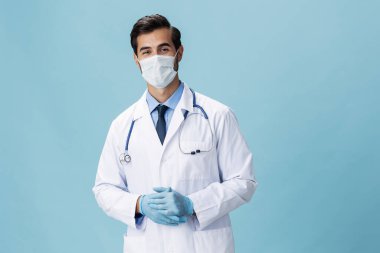 Beyaz önlüklü ve tıbbi maskeli bir erkek doktor elleriyle dikiliyor ve mavi izole bir arkaplanda kameraya bakıyor, kopyalama alanı, yazı için boşluk. Yüksek kalite fotoğraf