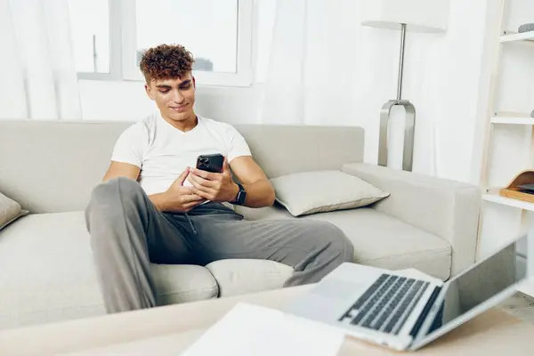 男子室内电话沙发成人微笑笔记本电脑自动通信坐智能手机卷曲使用智能拘留室家庭青少年现代 — 图库照片