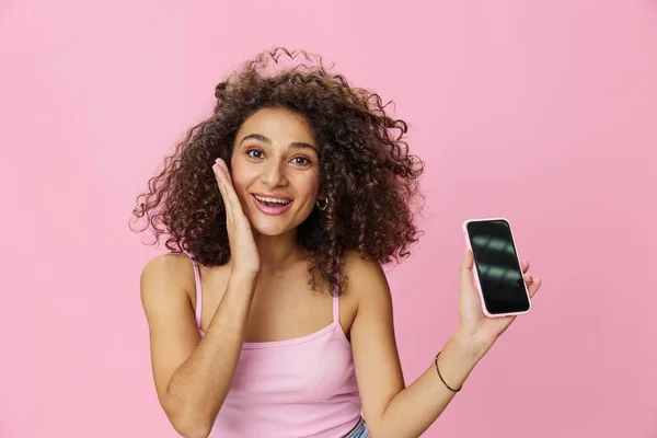 Woman blogger holding phone video call takes selfies, with curly hair in pink smile t-shirt and jeans poses on pink background, copy space, technology and social media, online. High quality photo