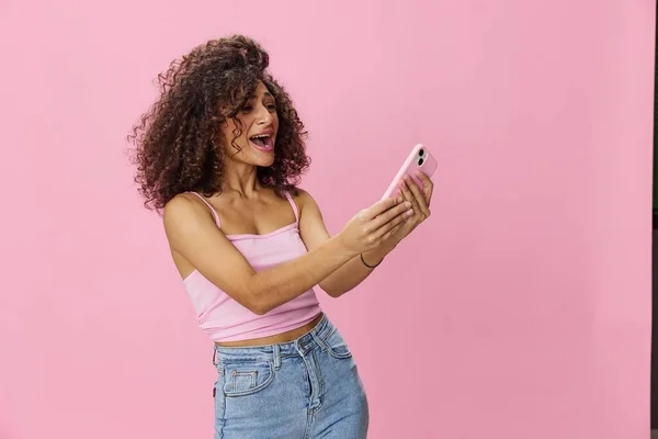 Woman blogger holding phone video call takes selfies, with curly hair in pink smile t-shirt and jeans poses on pink background, copy space, technology and social media, online. High quality photo