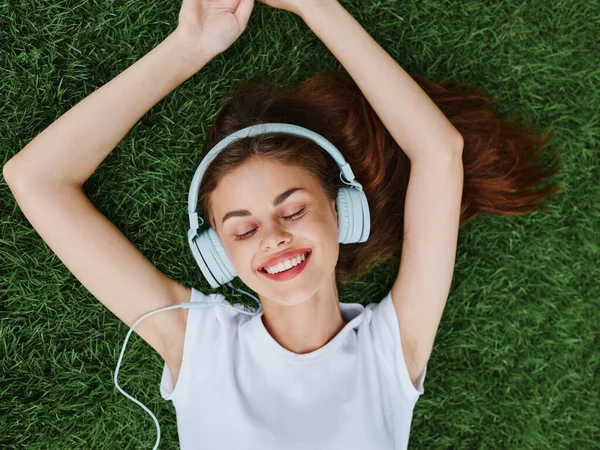 Woman wearing headphones lying on the green grass in a T-shirt and listening to music with a smile with teeth happiness summer vacations in nature in the park . High quality photo