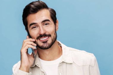 Esmer bir adamın portresi telefonda konuşurken gülümsüyor dişleri kıpır kıpır, kameraya bakıyor, mavi bir arka planda beyaz tişört ve kot pantolon, kopyalama alanı. Yüksek kalite fotoğraf