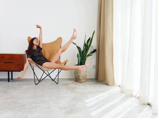 Woman sitting on a chair near the window smile cheerful feet and hands up, modern stylish interior Scandinavian lifestyle, copy space. High quality photo