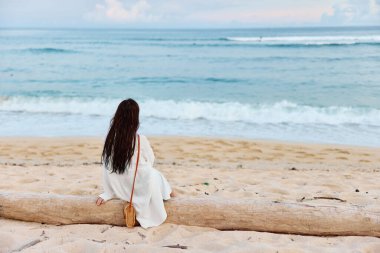 Yüzdükten sonra uzun ıslak saçlı bir kadın, sırtı kameraya dönük olarak kuma oturur ve Bali adasında okyanusun kenarında gün batımını seyreder. Yüksek kalite fotoğraf