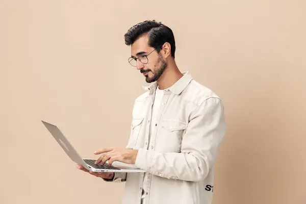 Portrait of a stylish man pensive look in glasses with a laptop in the hands of a freelancer, on a beige background in a white t-shirt, fashionable clothing style, space space. High quality photo