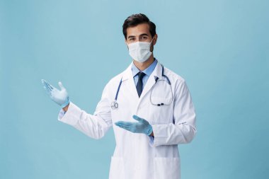 Beyaz önlüklü ve tıbbi maskeli erkek doktor el hareketlerini gösteriyor ve mavi izole arkaplandaki kameraya bakıyor, kopyalama alanı, yazı için boşluk. Yüksek kalite fotoğraf