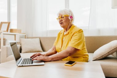 Kulaklıkla yazı yazan mutlu, yaşlı bir kadın. Sarı bir gömlekle evde oturuyor. Parlak, modern bir iç mekan. Yüksek kalite fotoğraf