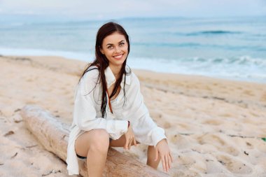 Beyaz mayo gömleği ve kot şortuyla gülümseyen mutlu bronz tenli kadın, Bali 'de yüzdükten sonra okyanusun kenarında ıslak saçlarıyla, gün batımı ışığı ve pembe bulutlarla oturuyor. Yüksek kalite fotoğraf