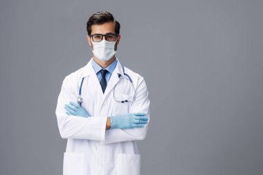 Beyaz önlüklü, tıbbi maskeli, steril eldivenli, gözlüklü bir erkek doktor gri izole bir arkaplanda kameraya bakar. Kopya alanı, yazı için yer, sağlık. Yüksek kalite fotoğraf