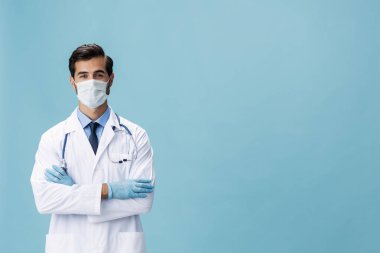 Beyaz önlüklü ve tıbbi maskeli erkek doktor el hareketlerini gösteriyor ve mavi izole arkaplandaki kameraya bakıyor, kopyalama alanı, yazı için boşluk. Yüksek kalite fotoğraf