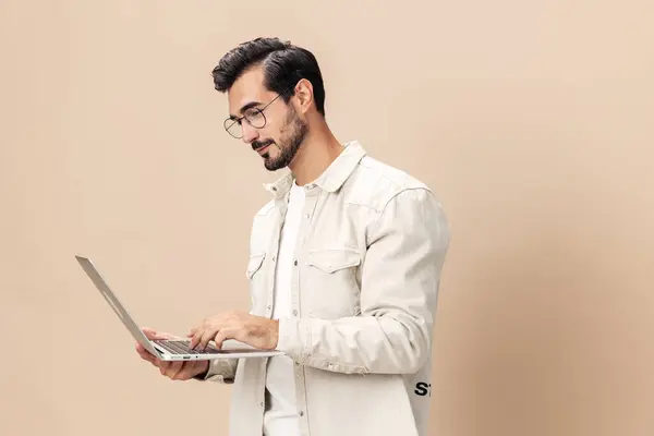 Portrait of a stylish man pensive look in glasses with a laptop in the hands of a freelancer, on a beige background in a white t-shirt, fashionable clothing style, space space. High quality photo